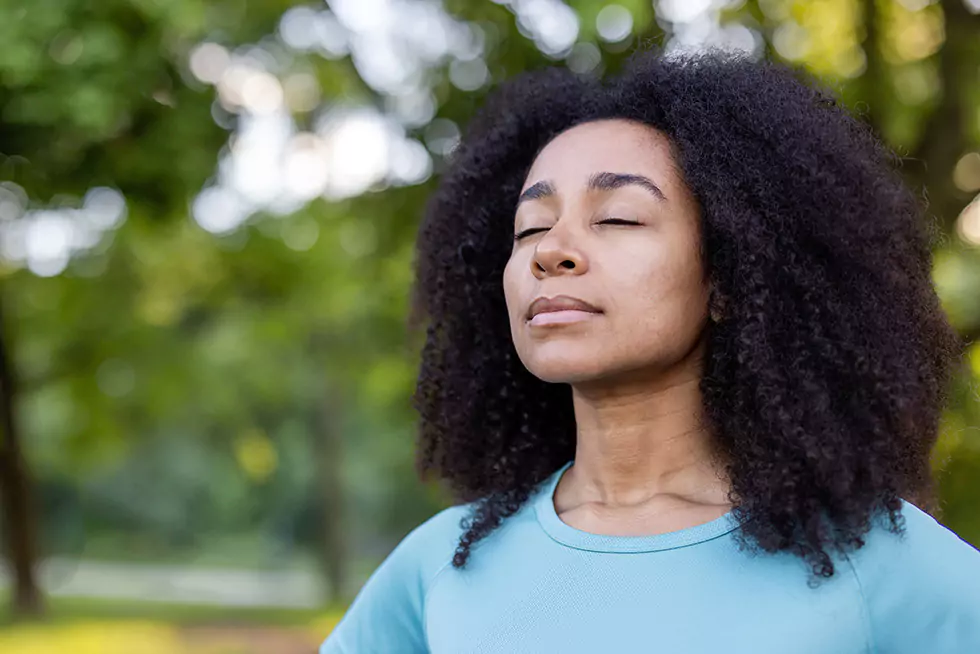 Frau praktiziert mit geschlossenen Augen achtsame Meditation in ruhiger Naturkulisse. Frau praktiziert achtsame Meditation in ruhiger Naturkulisse – mit geschlossenen Augen genießt sie den Moment der inneren Einkehr. Die Verbindung zur Natur fördert Entspannung, inneren Frieden und geistiges Wohlbefinden.
